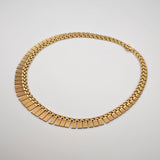 Goldene Halskette mit rechteckigen, gerillten Gliedern und feinen Kettensegmenten auf hellem Hintergrund.