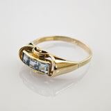 Goldring mit drei quadratischen hellblauen Edelsteinen in Goldener Ring mit rechteckigem Oberteil und drei blauen quadratischen Edelsteinen auf reflektierendem Untergrund.
