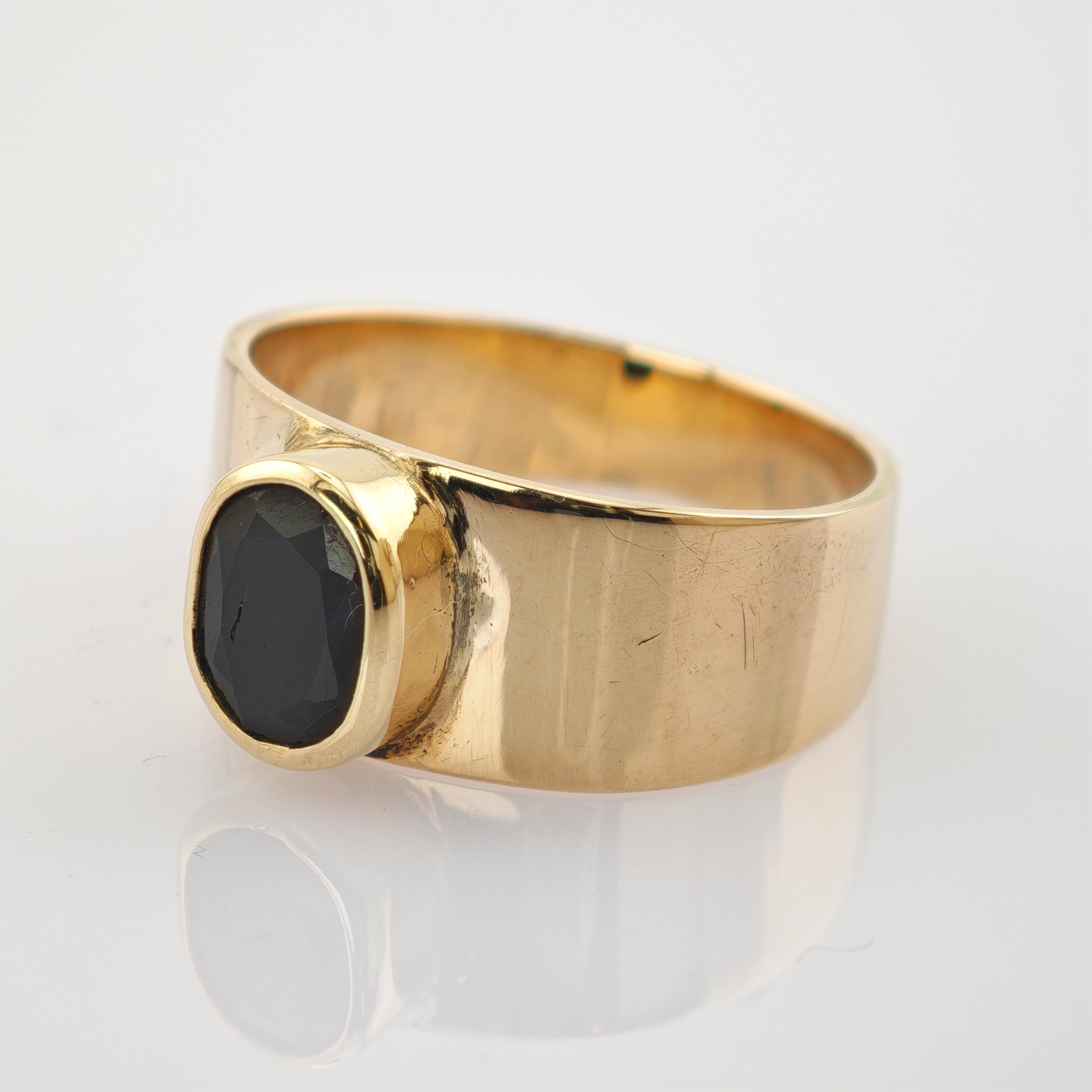Goldener Ring mit breitem Band und ovalem schwarzen Edelstein auf reflektierender Oberfläche