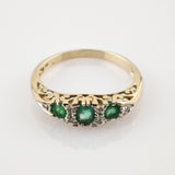 Goldener Ring mit filigraner Gravur und drei horizontal angeordneten Smaragden, umgeben von kleinen weißen Steinen.