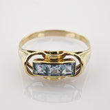 Goldener Ring mit drei quadratischen hellblauen Edelsteinen und geschwungenen Details auf weißem Goldener Ring mit drei quadratisch geschliffenen hellblauen Edelsteinen in Reihe