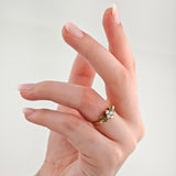 Hand mit ovalen, natürlich lackierten Nägeln und goldenem Ring mit Diamantcluster vor grauem Hintergrund