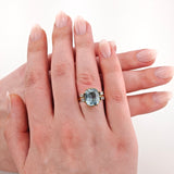Nahaufnahme einer linken Hand, die einen goldenen Ring mit einem großen kissenförmigen hellblauen Edelstein und kleinen Diamanten trägt.