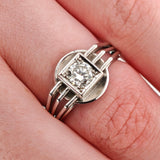 Nahaufnahme einer Hand mit silbernem Ring, geometrischem Design und Diamanten in quadratischem Rahmen.