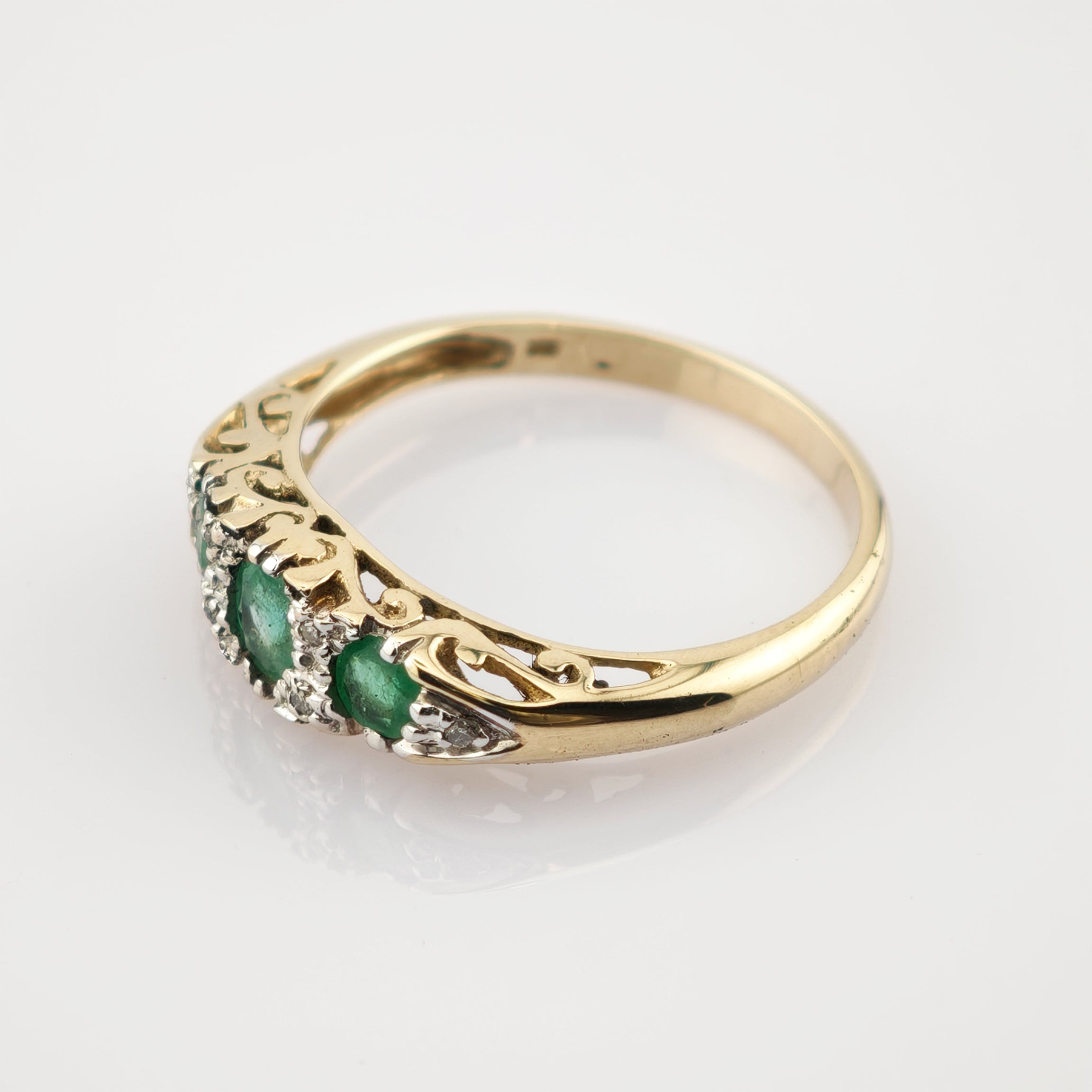 Goldener Ring mit filigraner Gravur und drei horizontal angeordneten Smaragden, umgeben von kleinen weißen Steinen.