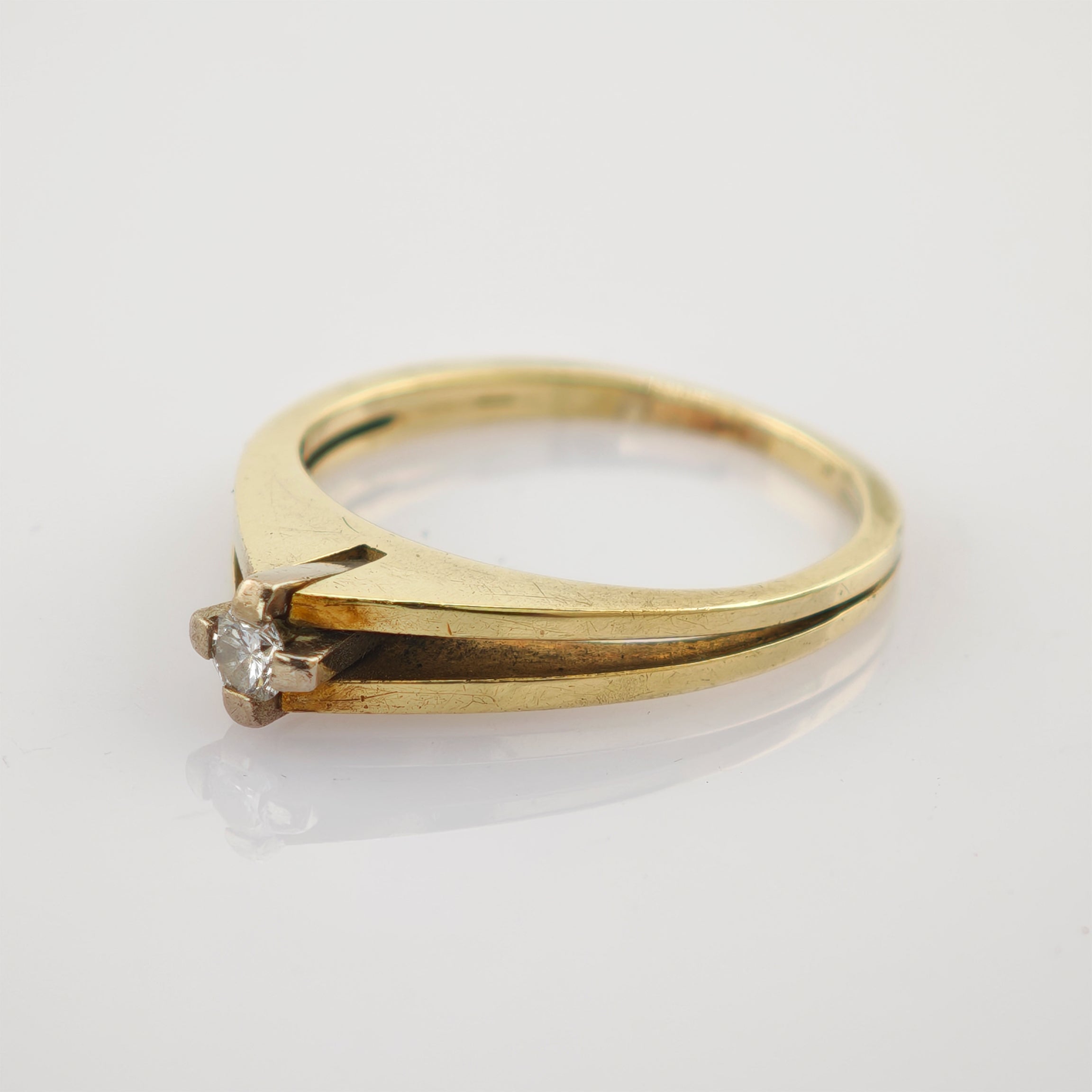 Schlichter Goldring mit schmalem Band und kleinem klarem Diamant in Vierkrappenfassung auf reflektierendem weißen Untergrund.
