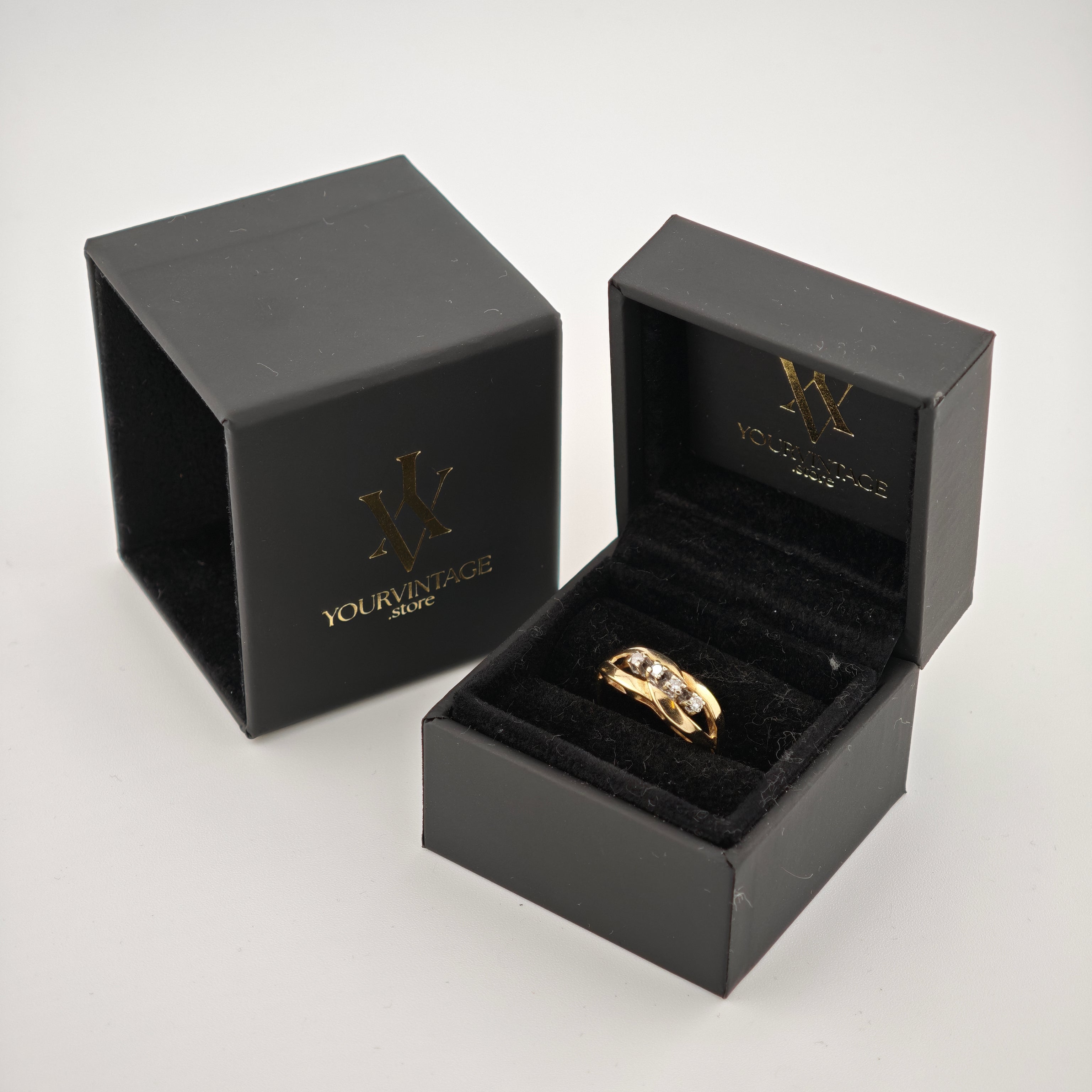 Schwarze Schmuckschachtel mit Goldring auf schwarzem Samt, daneben verzierte Außenschachtel mit Goldlogo auf weißem Hintergrund.