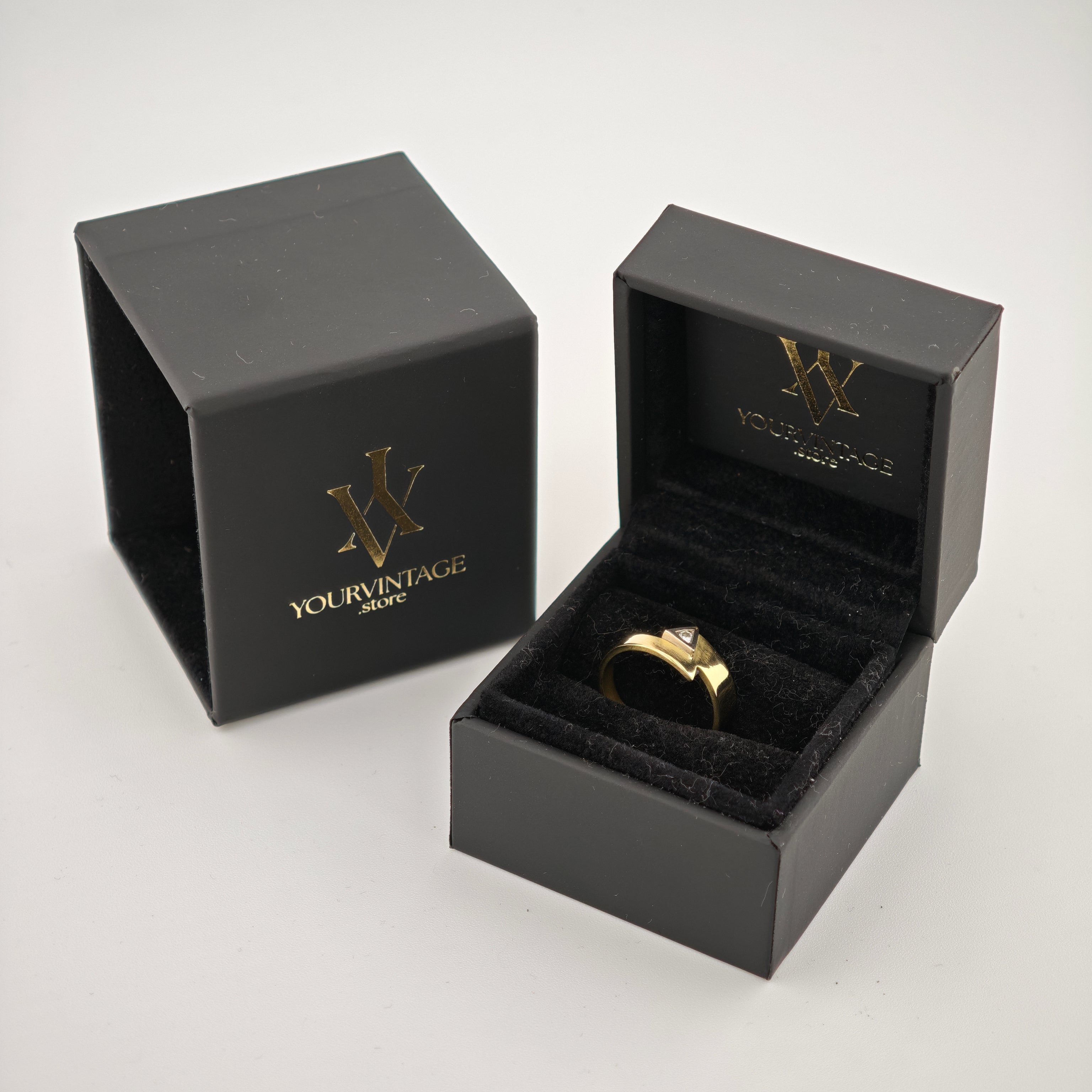 Zwei schwarze Schmuckkästchen mit goldenen Logos auf weißem Hintergrund, eines zeigt einen goldenen Ring mit dreieckigem Stein.