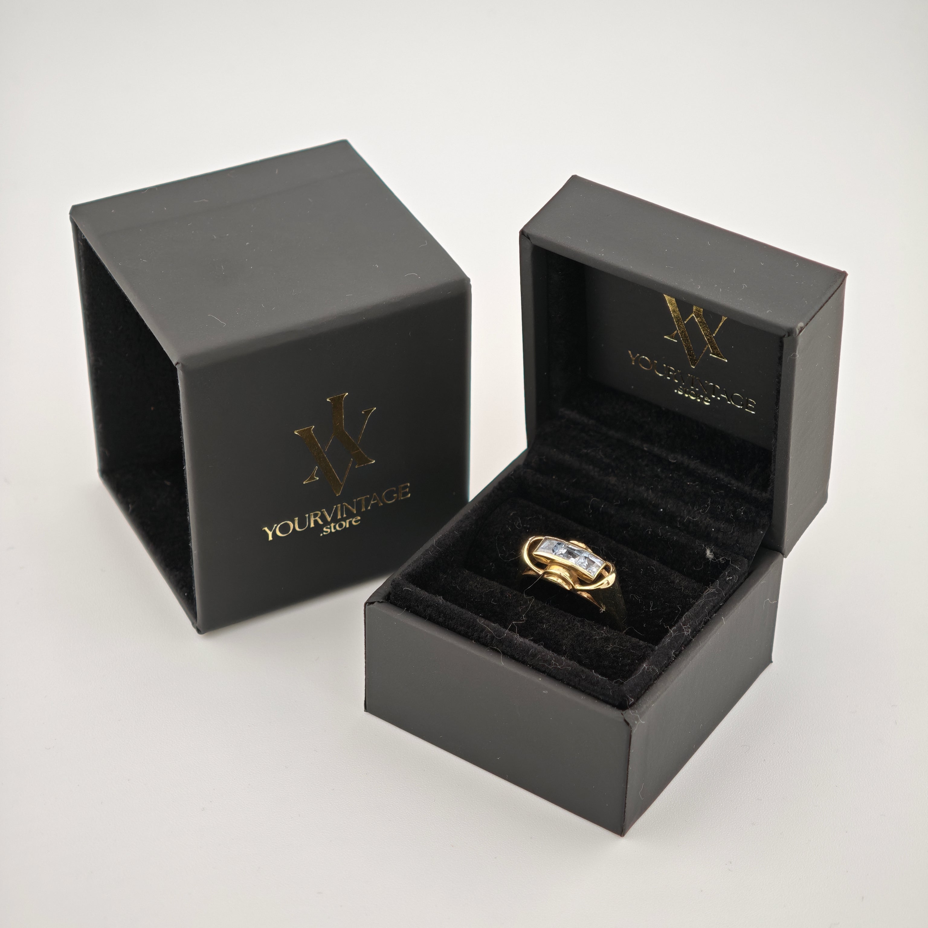 Schwarze Schmuckschachteln mit goldenem 'W'-Logo und Text 'yourvintoge.shop', offene Schachtel zeigt goldenen Ring mit drei klaren rechteckigen Edelsteinen.