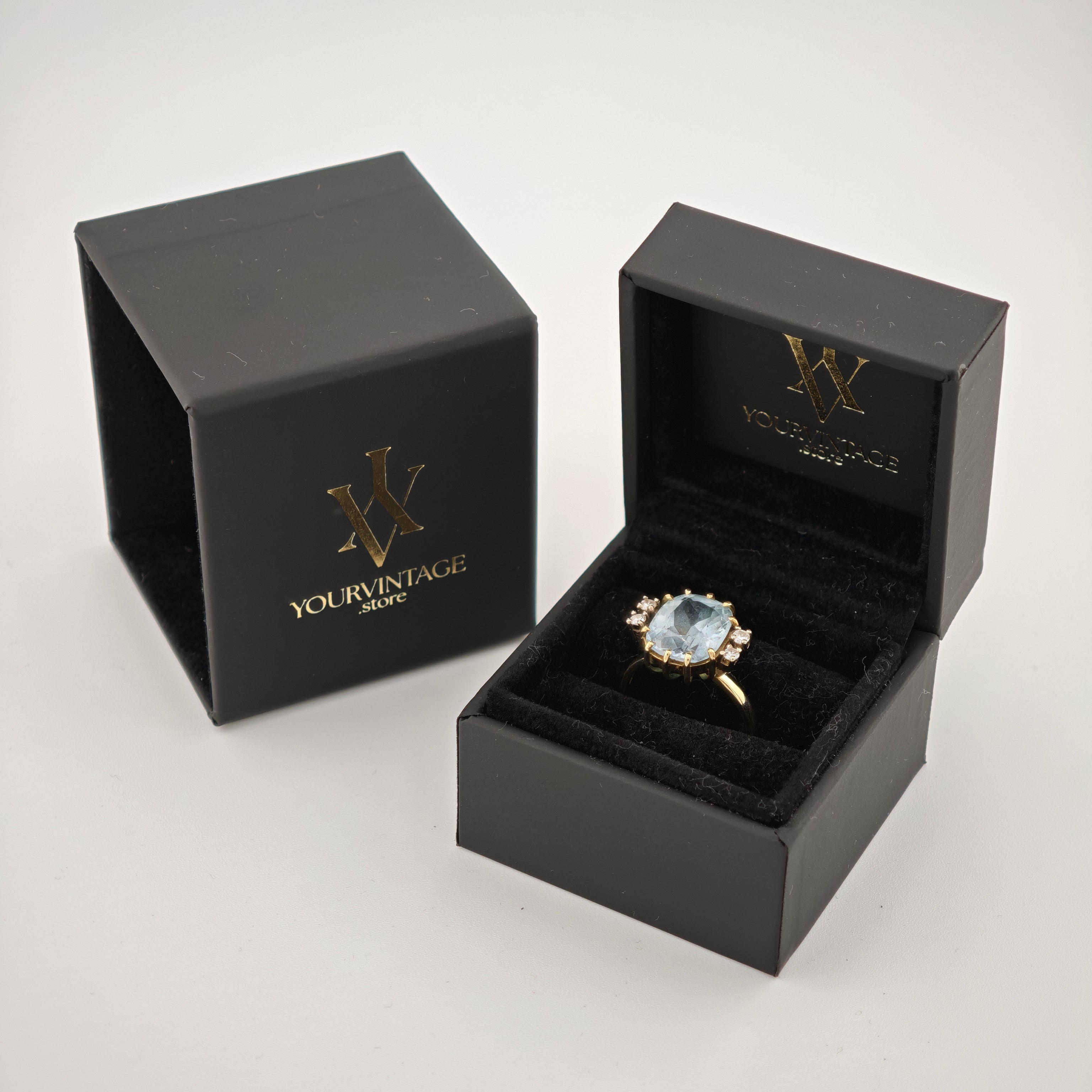 Zwei schwarze Schmuckschatullen, eine geschlossen mit goldener Aufschrift 'yourvintoge.shop', die andere offen mit einem Goldring und blauem Edelstein.