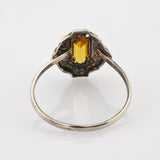 Silberner Ring mit rechteckigem gelbem Edelstein und verzierter Fassung auf reflektierender Oberfläche