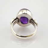 Silberner Ring mit großem ovalem violettem Edelstein und verzierten Bandseiten auf weißem Hintergrund.