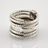 Nahaufnahme eines silbernen Rings mit verflochtenen, gedrehten Bändern und glattem Mittelband auf reflektierender Oberfläche.